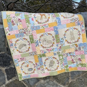 May include: A patchwork quilt with a whimsical woodland theme. The quilt features embroidered animal portraits, including a deer, rabbit, and owl, surrounded by floral wreaths. Pastel colors like pink, blue, and yellow are used throughout the design.