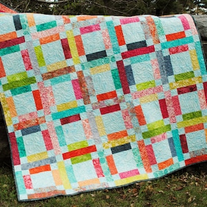 May include: A colorful quilt with a patchwork design featuring various shades of red, orange, yellow, green, blue, and turquoise. The quilt is made of fabric strips sewn together in a grid pattern. The quilt is hanging on a tree branch.