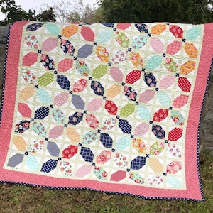 May include: A colorful quilt with a geometric pattern of overlapping diamonds and squares. The quilt is made with a variety of floral and geometric fabrics in shades of pink, red, blue, green, and yellow. The quilt is edged with a pink and white polka dot trim.