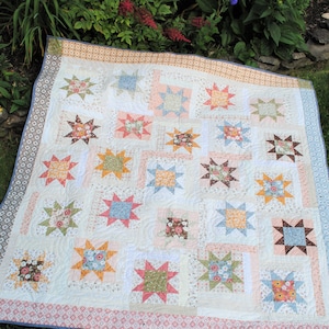 Op de afbeelding: Een wit quilt met een patchwork-ontwerp met verschillende bloemenstoffen in tinten roze, blauw, groen en bruin. Het quilt is gemaakt van vierkanten met stervormige patronen en is afgewerkt met een blauwe en witte rand.