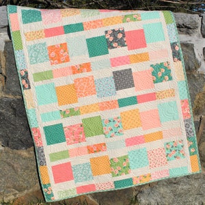 May include: A patchwork quilt featuring a mix of floral and geometric patterns in shades of pink, orange, green, and blue. The quilt is made up of squares and rectangles and is hanging on a stone wall.