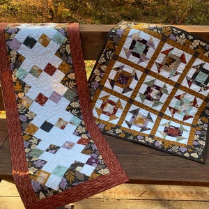 Könnte beinhalten: Zwei handgefertigte Quilts mit Herbstmustern. Einer ist ein Tischläufer mit weißer Mitte und Rautenmuster, umrandet von rostfarbenem Stoff. Der andere ist ein quadratischer Quilt mit einem geometrischen Design in Herbstfarben.