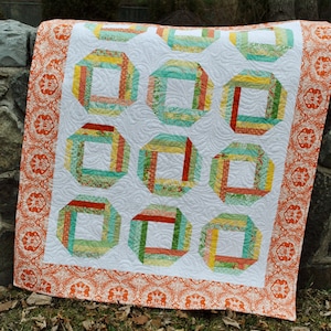 May include: A white quilt with a repeating pattern of orange, green, and yellow squares. The quilt is edged with a white and orange fabric with a floral pattern.