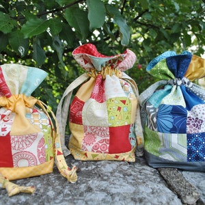 Peut inclure: Trois sacs &agrave; cordon de couleur faits de tissu patchwork. Les sacs sont rouge, jaune, bleu, vert et blanc. Les sacs sont pos&eacute;s sur une surface de pierre grise.