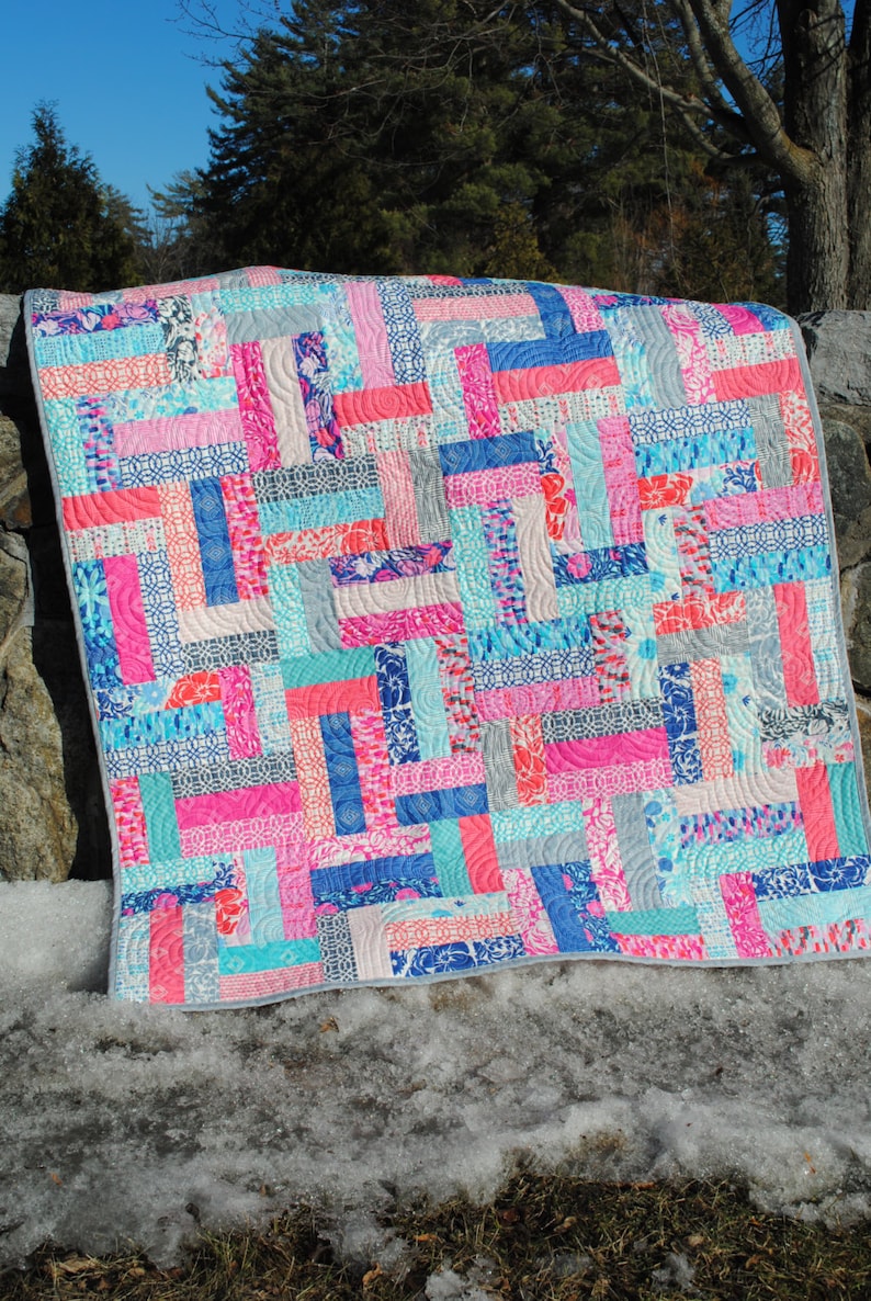 May include: A colorful quilt with a patchwork design featuring shades of pink, blue, and gray. The quilt is made of various fabrics with different patterns and textures. It is displayed on a snowy surface with green grass in the foreground.
