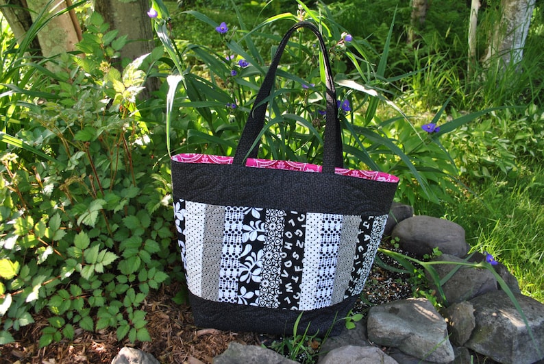 May include: A black and white patchwork tote bag with a pink lining. The bag has a black fabric handle and is made of different patterned fabrics.