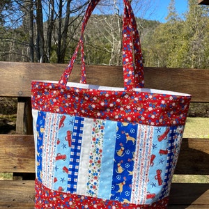 May include: Red, white, and blue fabric tote bag with a patchwork design featuring stars, stripes, and a patriotic theme. The bag has a large main compartment and two long handles.