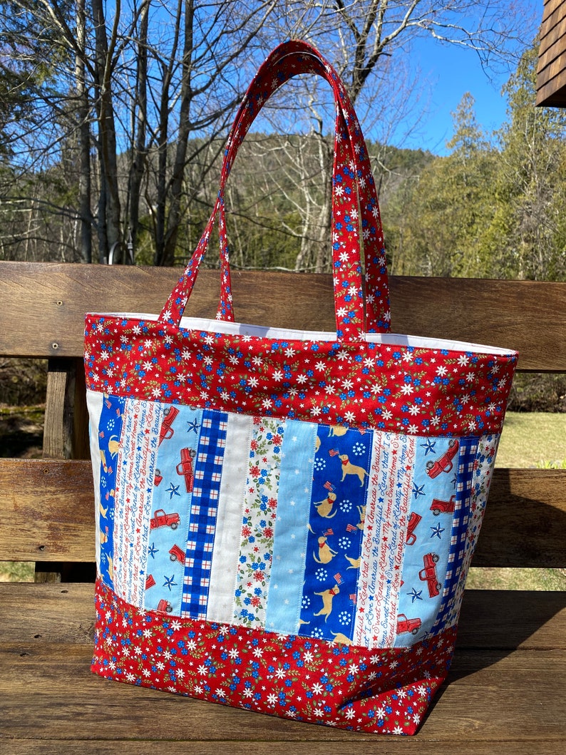 May include: Red, white, and blue fabric tote bag with a patchwork design featuring stars, stripes, and a patriotic theme. The bag has a large main compartment and two long handles.