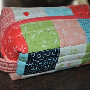 Peut inclure: Une pochette zippée en tissu patchwork rose et vert avec un motif géométrique noir et blanc. La pochette a une anse en tissu rose.