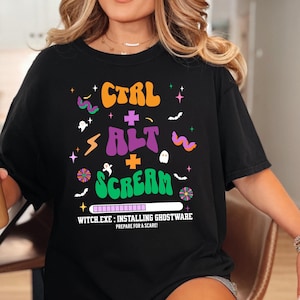 以下が含まれることがあります： 黒いTシャツ。ハロウィーンをテーマにしたカラフルなグラフィックデザイン。オレンジ、紫、緑で「CTRL ALT SCREAM」の文字、ハロウィーンのアイコン、そして「WITCH.EXE: INSTALLING GHOSTWARE. PREPARE FOR A SCARE!」のテキストが含まれています。