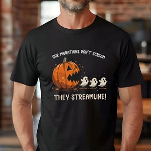 May include: Black t-shirt featuring a Halloween-themed graphic. The design includes an orange jack-o'-lantern with a jagged mouth and three white ghosts. Text above reads "OUR MIGRATIONS DON'T SCREAM" and below "THEY STREAMLINE!"