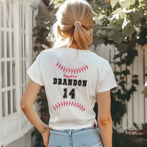 以下が含まれることがあります： 後ろにピンクの野球のグラフィックと「Biggest fan BRANDON 14」の文字が入った白いTシャツです。