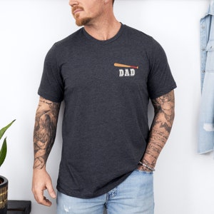 Könnte beinhalten: Ein dunkelgraues T-Shirt mit einem Baseballschläger-Motiv und dem Wort "DAD" auf der Vorderseite.