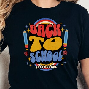 Puede incluir: Camiseta negra con un gráfico de estilo retro que dice "Back to School Third Grade" en letras de colores. El gráfico incluye un arcoíris, estrellas y un lápiz.