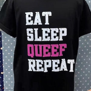 May include: Black t-shirt with the words "EAT SLEEP QUEEF REPEAT" in white and pink block letters. The shirt has a crew neck and short sleeves. The text is centered on the front of the shirt.