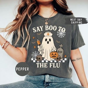 May include: A charcoal gray t-shirt featuring a graphic of a ghost dressed as a nurse holding a syringe and a jack-o'-lantern. The design includes skulls, spiderwebs, a tombstone, and the text "SAY BOO TO THE FLU" in a bold, cream-colored font.