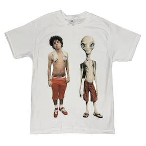 May include: White t-shirt featuring a graphic of a person and an alien. The person is wearing orange pants and red shoes. The alien is wearing red shorts and a backpack. The t-shirt is a casual style.