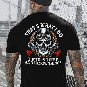 May include: Black t-shirt featuring a skull wearing a motorcycle helmet and goggles, with wrenches and flames. The text reads "THAT'S WHAT I DO, I FIX STUFF AND I KNOW THINGS."