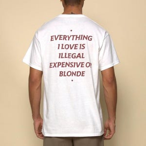 May include: White t-shirt with the text "EVERYTHING I LOVE IS ILLEGAL EXPENSIVE OR BLONDE" in burgundy lettering. The shirt is a crew neck and has short sleeves.