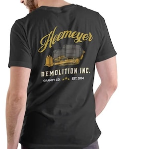 May include: Black t-shirt with a gold and yellow graphic of a bulldozer and the text "Heemeyer Demolition Inc. Granby Co. Est. 2004". The shirt is made of a soft material.
