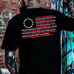 May include: A black t-shirt featuring a red, distressed American flag graphic on the back. The design includes a circle of thirteen white stars and the text: We hold these truths to be self evident, that all men are created equal.