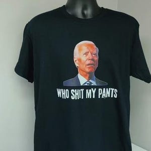 May include: Black t-shirt with a graphic of a man's face and the text "WHO SHIT MY PANTS". The words "SLEEPY JOE" are in yellow letters above the shirt. The shirt is displayed on a black mannequin.