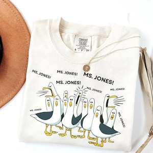 May include: Cream-colored Comfort Colors t-shirt with a graphic of cartoon seagulls and the text "MS. JONES!". The seagulls are white with gray wings and yellow feet. A gold necklace is draped on the shirt.