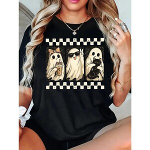 May include: A black short-sleeve t-shirt featuring a graphic print of three white ghosts arranged in a row. The ghosts are framed by black and white checkered borders. One ghost wears a bow, one wears sunglasses, and one holds a black cat.