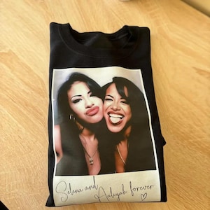 May include: Black t-shirt featuring a photo of Selena and Aaliyah. The photo is in a white frame and includes the text "Selena and Aaliyah forever" in cursive script. The image shows the two women smiling.