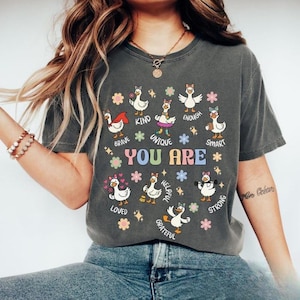May include: Dark gray t-shirt featuring a colorful graphic of cartoon ducks with positive affirmations. The text "YOU ARE" is prominently displayed, surrounded by words like "Brave," "Kind," and "Smart." The design includes stars and floral accents.