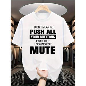May include: White t-shirt with the text "I DIDN'T MEAN TO PUSH ALL YOUR BUTTONS I WAS JUST LOOKING FOR MUTE" in black. The shirt is displayed on a hanger against a neutral background.