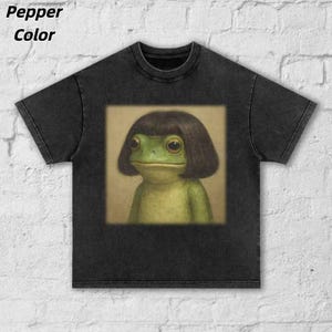 May include: Pepper color t-shirt featuring a whimsical illustration of a frog with a bob haircut. The frog is green with large eyes, set against a neutral background. The shirt is a dark gray color.