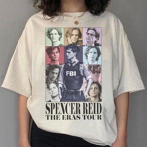May include: A beige t-shirt with a graphic print of a collage of different portraits of a man. The text "Spencer Reid The Eras Tour" is printed below the collage.
