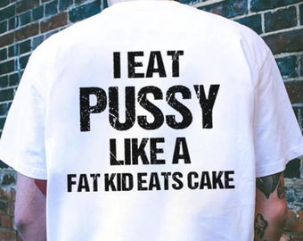 May include: White t-shirt with the text "I EAT PUSSY LIKE A FAT KID EATS CAKE" printed in black. The shirt is being worn in front of a brick wall.