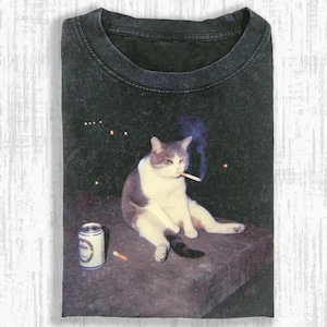 May include: Dark gray t-shirt featuring a cat smoking a cigarette, with a beer can and a cigarette butt. The cat is white and gray, sitting on a stone surface against a dark background with lights.