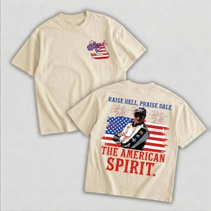 May include: Cream-colored t-shirts with patriotic designs. One shirt features a red, white, and blue number 3 logo. The other shirt displays an image of a man in front of an American flag with the text "RAISE HELL, PRAISE DALE" and "THE AMERICAN SPIRIT."