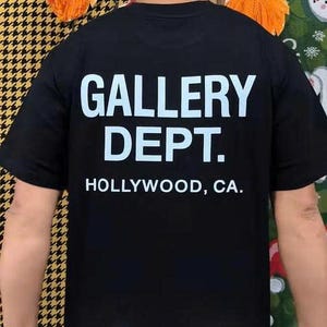 May include: Black t-shirt with the text "GALLERY DEPT." in large white block letters. Below the text is "HOLLYWOOD, CA." in smaller white letters. The shirt is short-sleeved.