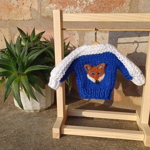 May include: A blue knitted sweater with white trim and a fox face design. The sweater is hanging on a wooden clothes rack.