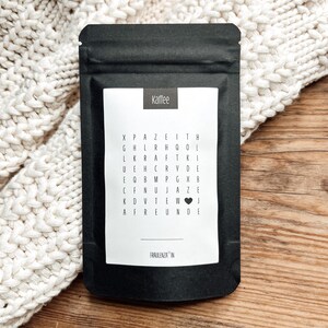 Könnte beinhalten: Ein schwarzer Kaffeebeutel mit einem weißen Etikett mit dem Wort "Kaffee" und einem Wortsuchrätsel. Das Rätsel enthält Wörter wie "Zeit" und "Freunde". Der Beutel ist versiegelt und liegt auf einer Holzoberfläche.