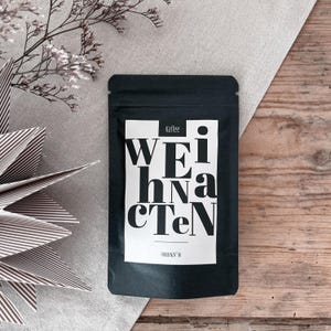 Könnte beinhalten: Ein schwarzer Kaffeebeutel mit dem Wort "Weihnachten" in großen weißen Buchstaben. Das Wort "Kaffee" steht über dem Haupttext. Der Beutel liegt auf einer Holzoberfläche mit dekorativen Elementen.