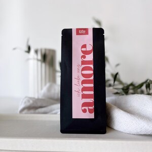 Könnte beinhalten: Ein schwarzer Kaffeebeutel mit einem rosa Etikett mit dem Wort "amore" in roter Schrift. Das Etikett enthält auch den deutschen Satz "ich liebe uns" und das Wort "Kaffee". Der Beutel wird auf einer weißen Oberfläche präsentiert.