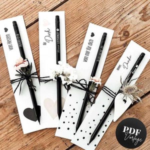 Peut inclure: Quatre stylos noirs avec des manchons en papier blanc, chacun orné de fleurs et de texte. Les stylos sont disposés sur une surface en bois. Le texte sur les manchons comprend des phrases comme "Von Herzen" et "Danke."