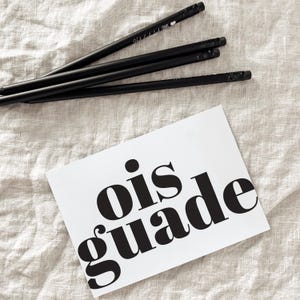 Könnte beinhalten: Vier schwarze Bleistifte mit Radiergummis und weißem Text sind neben einer weißen Karte mit den Worten "ois guade" in fetter schwarzer Schrift angeordnet. Die Artikel liegen auf einer strukturierten, hellfarbigen Oberfläche.