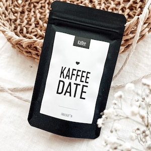 Könnte beinhalten: Ein schwarzer Kaffebeutel mit einem weißen Etikett, auf dem "Kaffee Date" mit einem kleinen Herzen über dem Text steht. Das Etikett trägt auch den Schriftzug "Frauenlenza" am unteren Rand.
