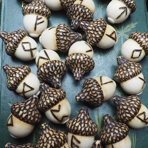 May include: A collection of 25 wooden acorns with different runes carved into them. The acorns are arranged on a green book with a black and white patterned background.