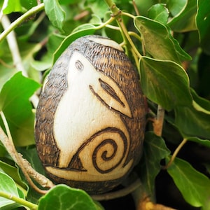 May include: A wooden egg with a carved rabbit design. The egg is brown and the rabbit is white. The egg is surrounded by green leaves.