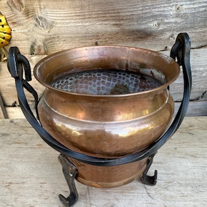 May include: A round copper pot with a hammered interior texture, featuring a wide opening and a smooth exterior finish. The pot is supported by a black metal stand with three curved legs and two side handles attached to the frame.