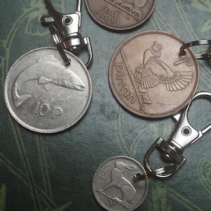 Puede incluir: Llaveros con monedas antiguas con varios diseños, como un pez, un pájaro y un hipopótamo. Cada moneda está unida a un cierre metálico plateado. Los llaveros están colocados sobre una superficie verde oscuro con patrones.