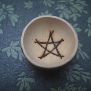 May include: A small, round, light-colored wooden bowl featuring a dark, hand-burned pentagram symbol centered at the bottom of the interior. The bowl has smooth, curved walls and a natural wood grain finish, presenting a simple, circular design.