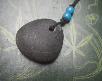 May include: A dark gray, smooth stone pendant with a drilled hole, strung on a black cord with two turquoise beads. The stone has subtle white markings. The background is a patterned green surface.